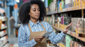 Jovem mulher afrodescendente, sorrindo levemente, utilizando um tablet enquanto trabalha em frente a uma gôndola de supermercado repleta de produtos. Ela veste um uniforme profissional e está focada em suas tarefas, demonstrando eficiência e dedicação.